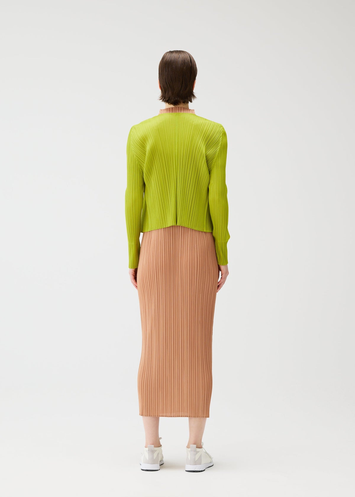 A person wears the PLEATS PLEASE ISSEY MIYAKE "MONTHLY COLORS : JUNE" collection—a lime green long-sleeve top with a peach pleated midi skirt. They have short brown hair, white sneakers, and are set against a plain white background. view 9 of 9