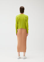 A person wears the PLEATS PLEASE ISSEY MIYAKE "MONTHLY COLORS : JUNE" collection—a lime green long-sleeve top with a peach pleated midi skirt. They have short brown hair, white sneakers, and are set against a plain white background.