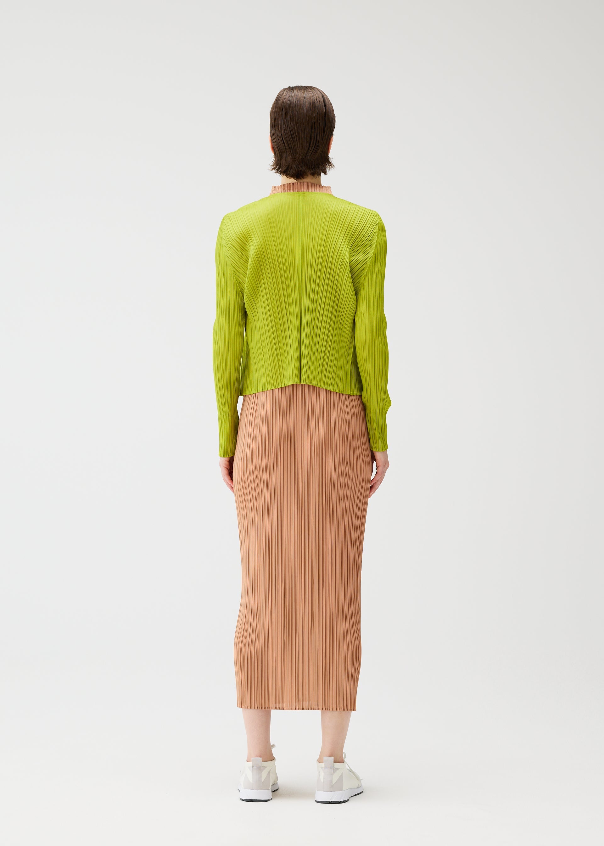 Zoomed view A person wears the PLEATS PLEASE ISSEY MIYAKE "MONTHLY COLORS : JUNE" collection—a lime green long-sleeve top with a peach pleated midi skirt. They have short brown hair, white sneakers, and are set against a plain white background.