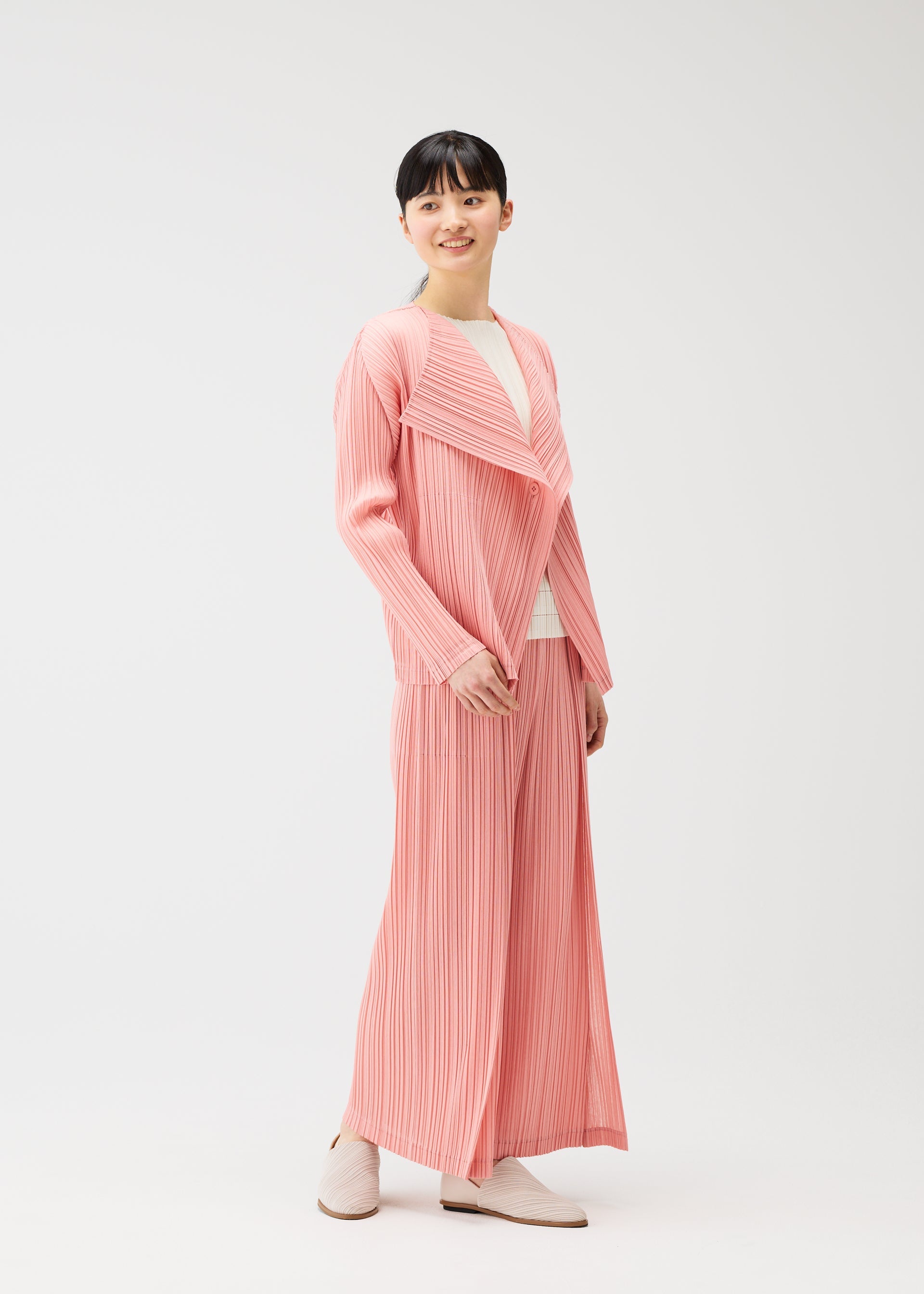 Zoomed view A person smiles in the RAMIE PLEATS outfit by PLEATS PLEASE ISSEY MIYAKE: a light pink pleated long jacket, wide-legged pants, and a white top, paired with light slip-on shoes against a plain white background.