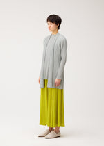A person with short dark hair wears a light gray MELLOW PLEATS long-sleeve top and bright yellow pleated skirt from PLEATS PLEASE ISSEY MIYAKE, styled with white slip-on shoes, standing against a plain white background.