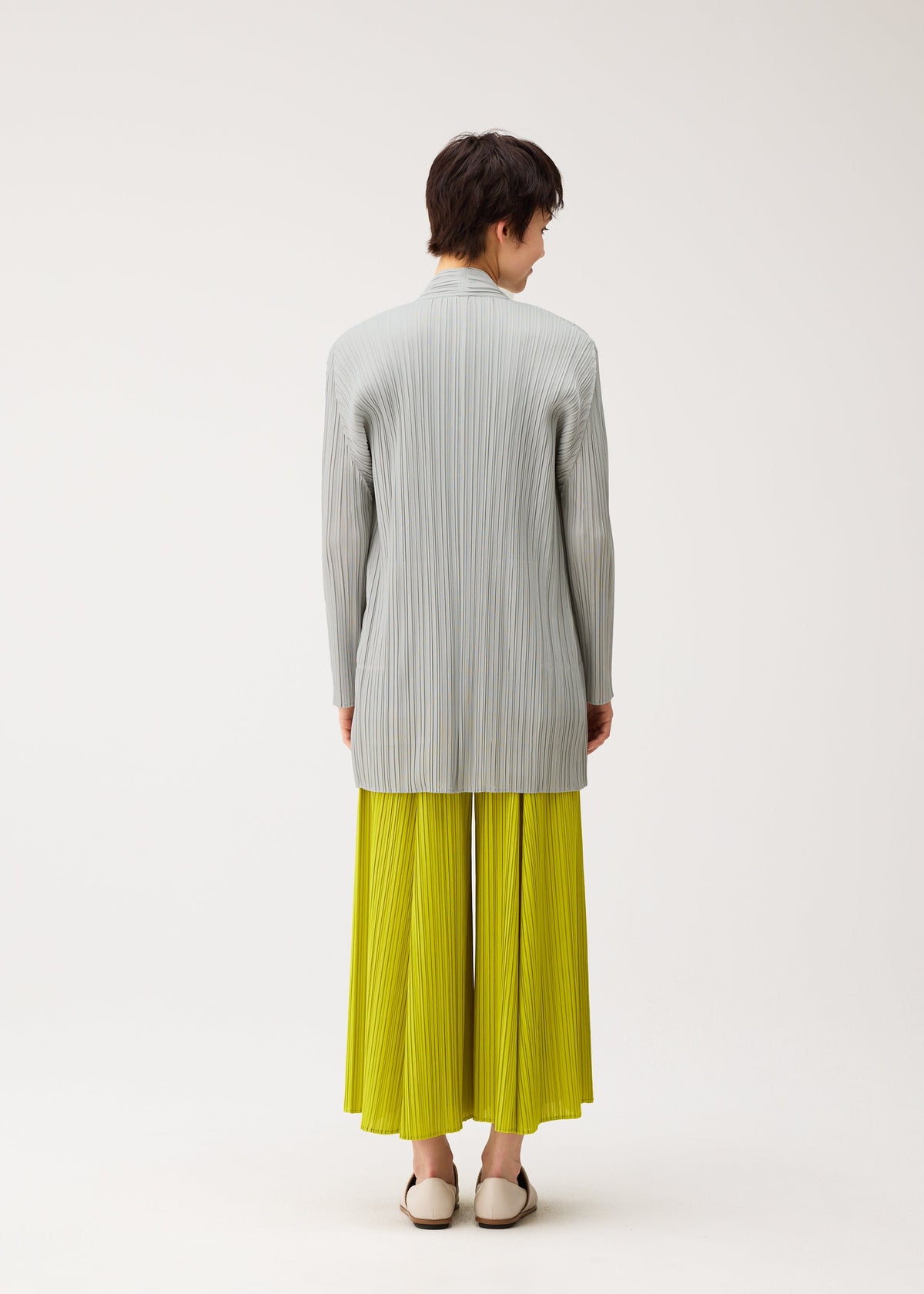 A person stands against a light background, wearing the MELLOW PLEATS bright yellow pleated skirt by PLEATS PLEASE ISSEY MIYAKE, paired with a light gray soft-textured mid-length cardigan and white shoes. view 8 of 9