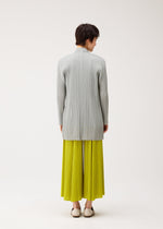A person stands against a light background, wearing the MELLOW PLEATS bright yellow pleated skirt by PLEATS PLEASE ISSEY MIYAKE, paired with a light gray soft-textured mid-length cardigan and white shoes.