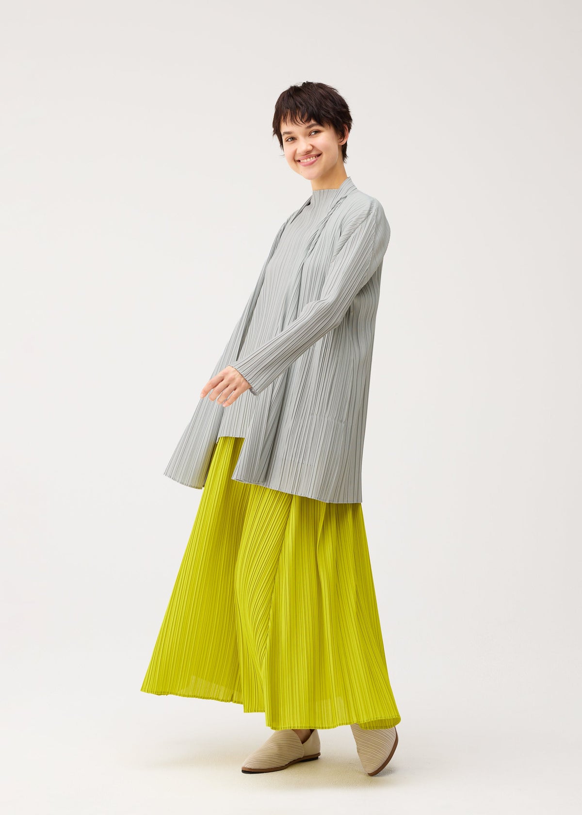 A person with short dark hair smiles in PLEATS PLEASE ISSEY MIYAKE's MELLOW PLEATS: a loose, light gray pleated top and bright yellow pleated wide-leg pants, paired with beige shoes against a plain white background. view 9 of 9