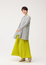 A person with short dark hair smiles in PLEATS PLEASE ISSEY MIYAKE's MELLOW PLEATS: a loose, light gray pleated top and bright yellow pleated wide-leg pants, paired with beige shoes against a plain white background.