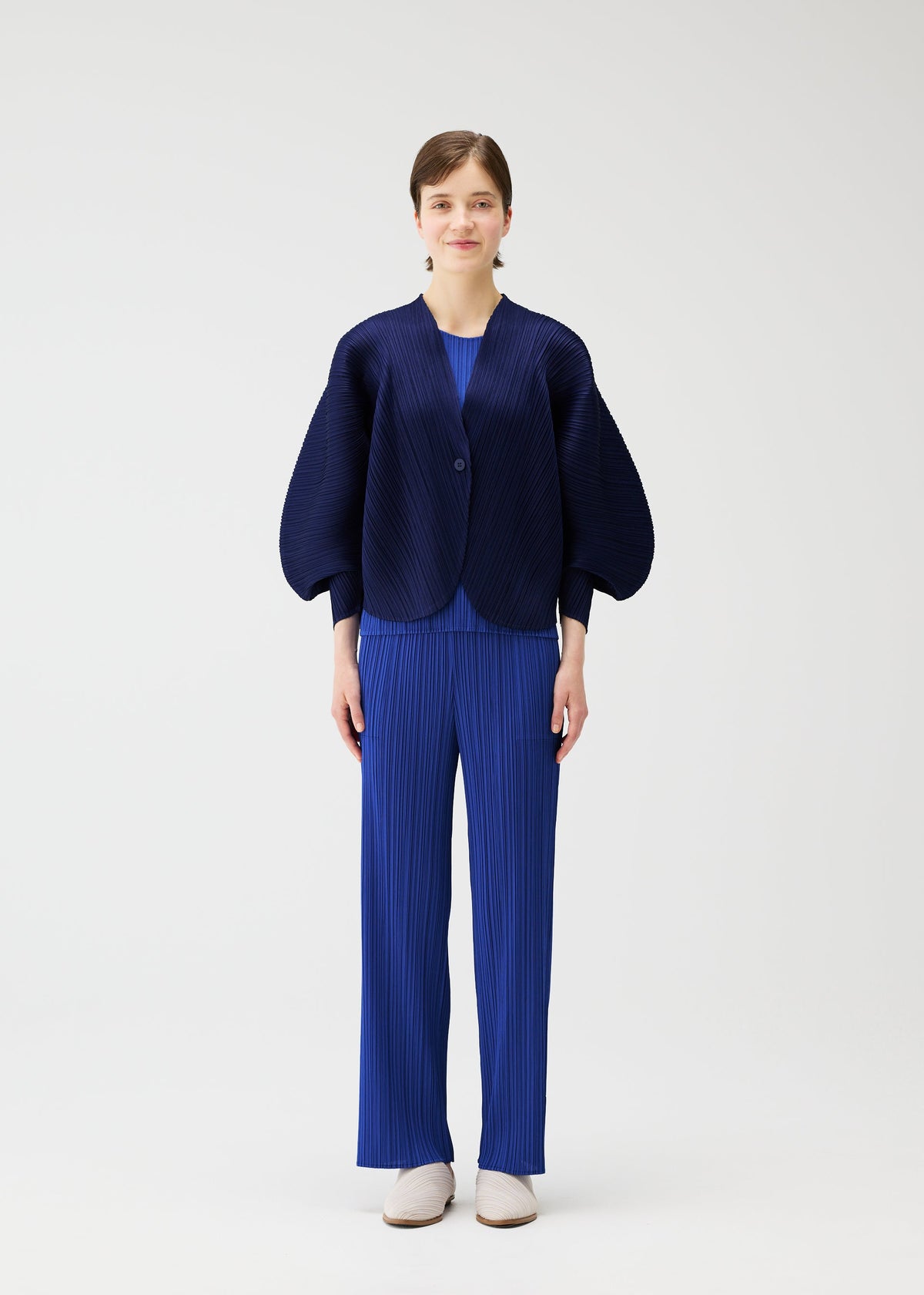 A person stands against a white background wearing the COAST pleated jacket and pants by PLEATS PLEASE ISSEY MIYAKE, featuring wide sleeves, an airy silhouette, short hair, and white shoes. view 5 of 7
