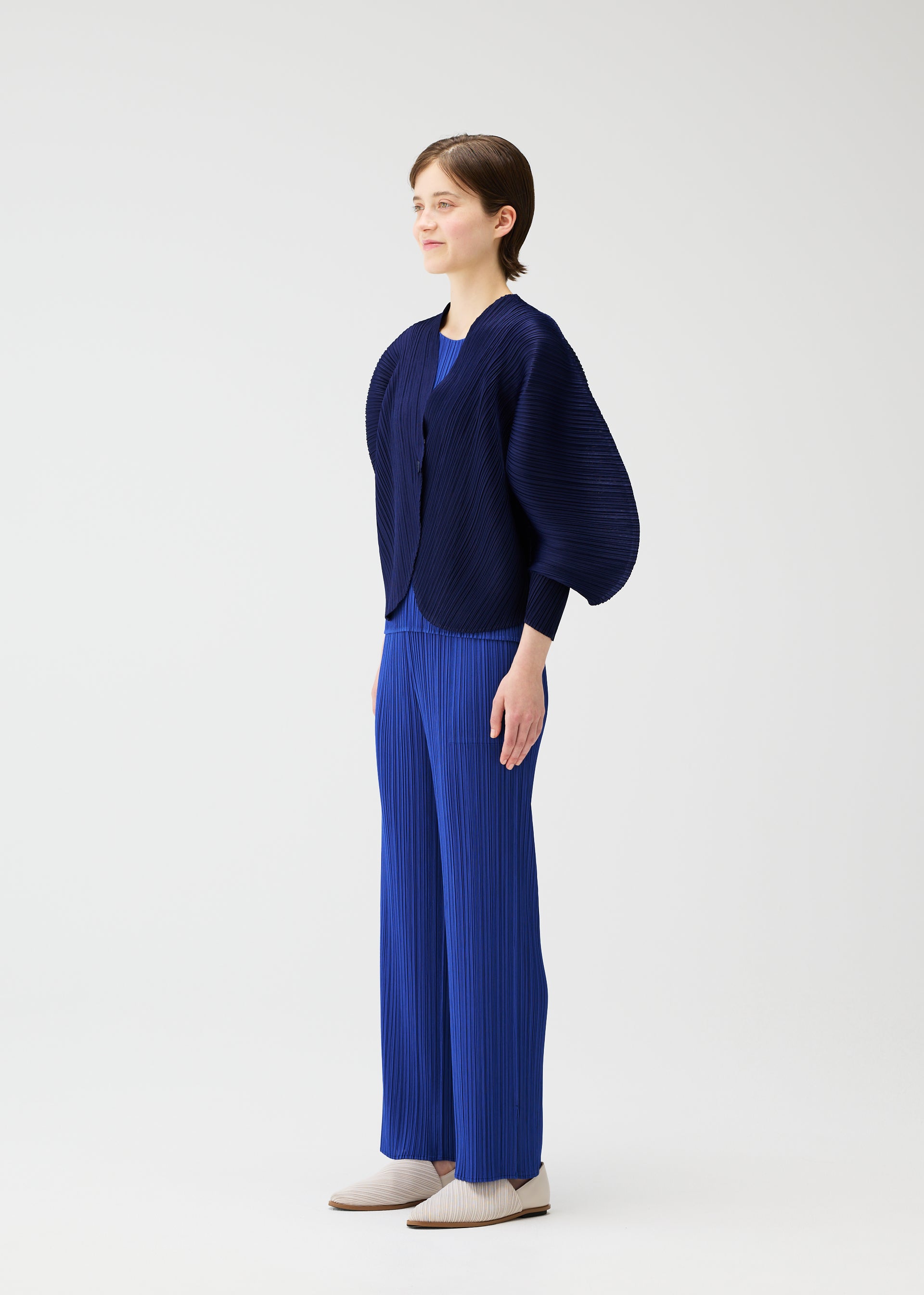 Zoomed view A person wears the COAST by PLEATS PLEASE ISSEY MIYAKE: a dark blue textured jacket with an airy silhouette over a bright blue top and matching pleated pants, paired with light shoes against a plain white background.