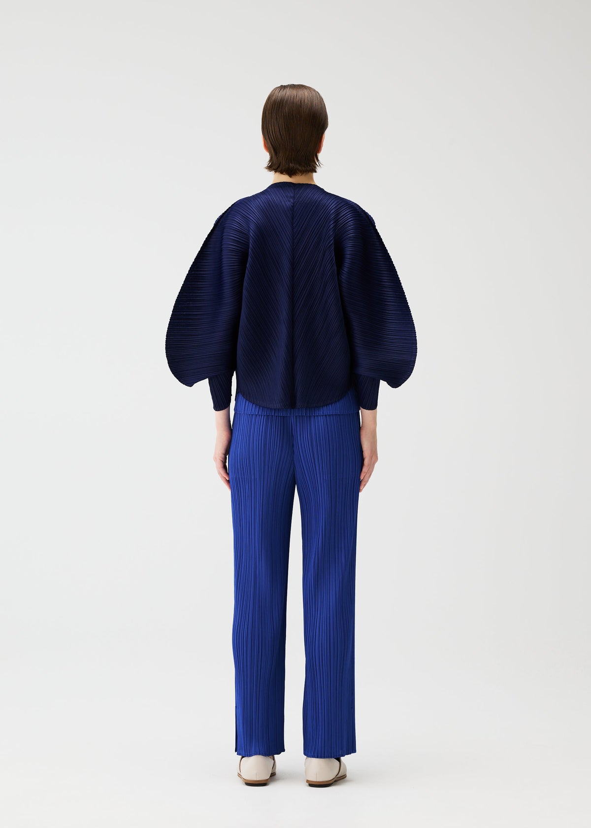 A person with short hair, seen from behind, wears the COAST jacket and bright blue pleated pants from PLEATS PLEASE ISSEY MIYAKE. The outfit features an airy silhouette and wide sleeves, styled with white shoes against a plain white background. view 7 of 7
