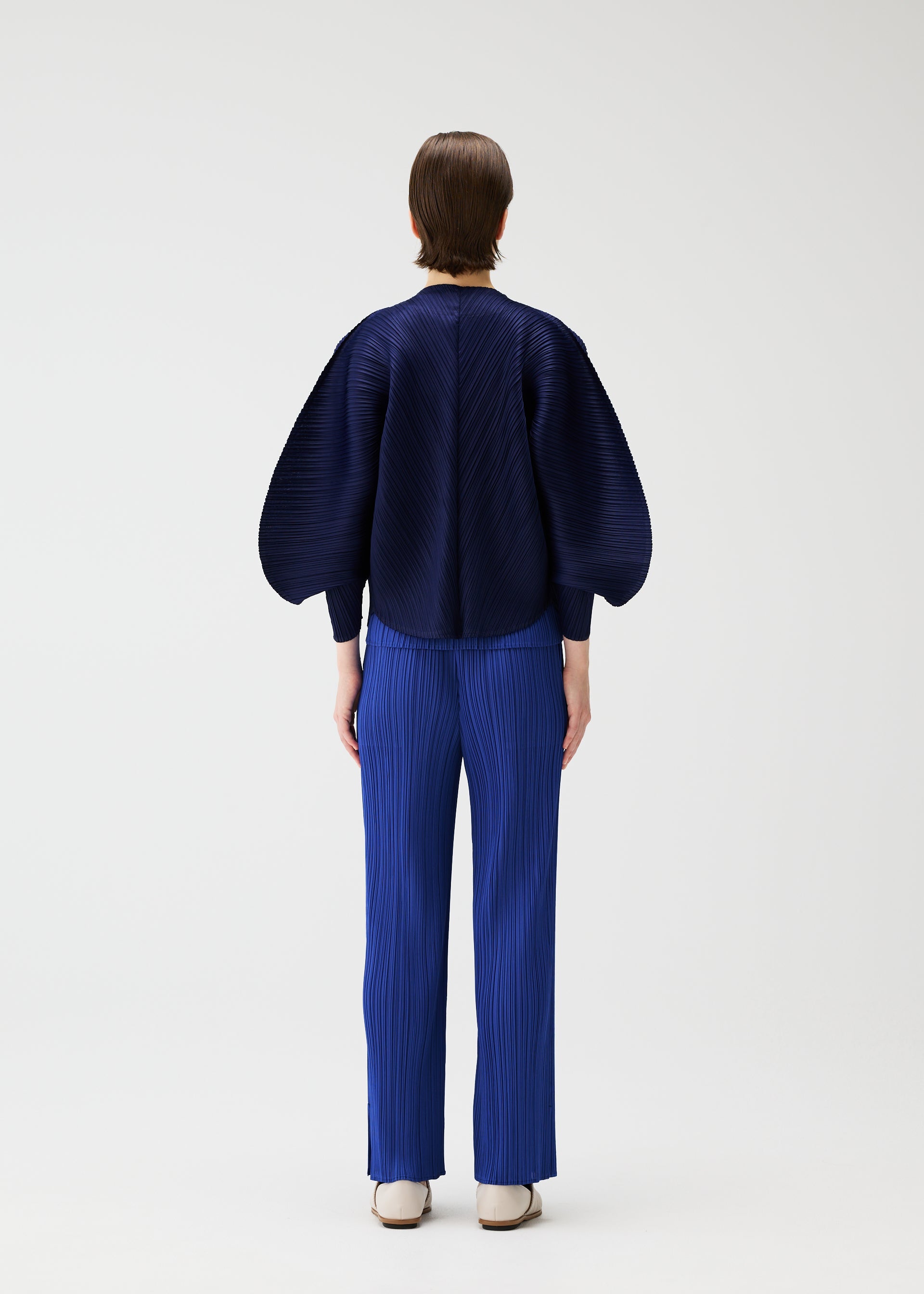 Zoomed view A person with short hair, seen from behind, wears the COAST jacket and bright blue pleated pants from PLEATS PLEASE ISSEY MIYAKE. The outfit features an airy silhouette and wide sleeves, styled with white shoes against a plain white background.
