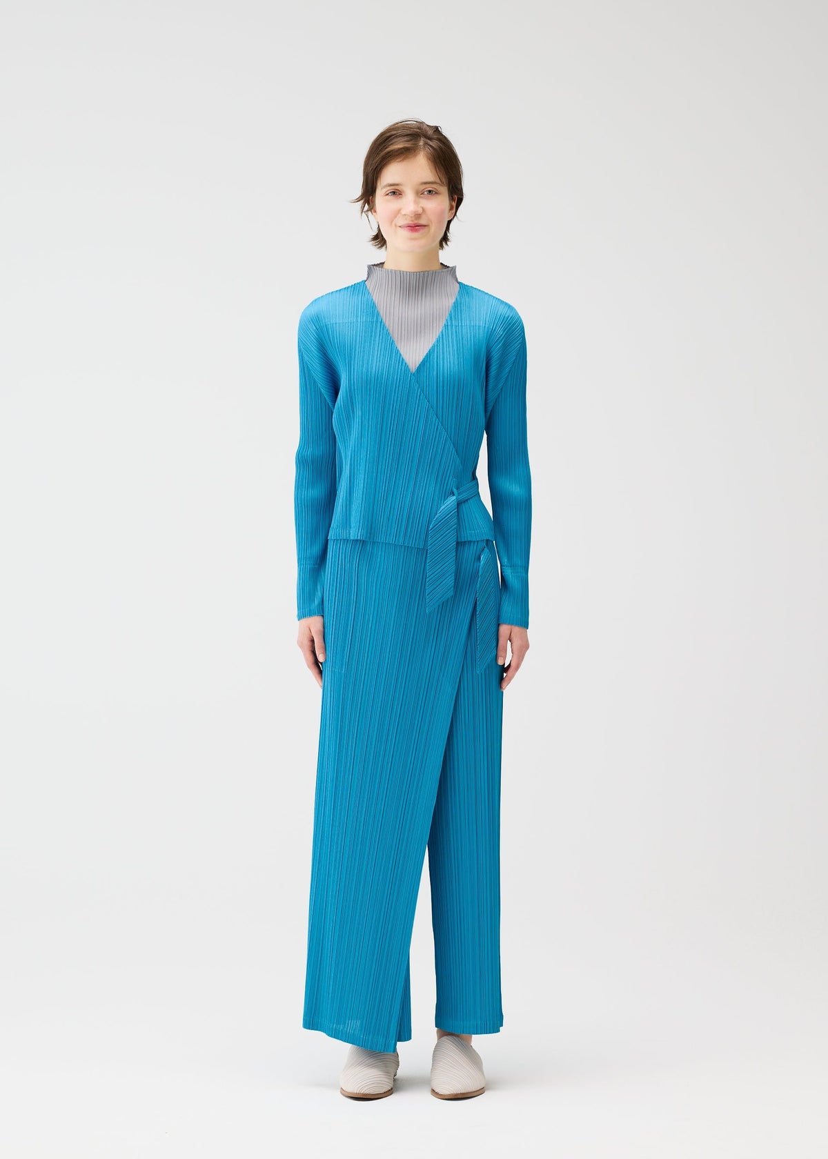 A person stands against a plain white background wearing the FACADE set by PLEATS PLEASE ISSEY MIYAKE: a bright blue long-sleeve wrap top with adjustable cord, matching wide-leg pants over a light gray turtleneck, and light-colored shoes. view 6 of 9