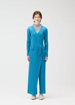 A person stands against a plain white background wearing the FACADE set by PLEATS PLEASE ISSEY MIYAKE: a bright blue long-sleeve wrap top with adjustable cord, matching wide-leg pants over a light gray turtleneck, and light-colored shoes.