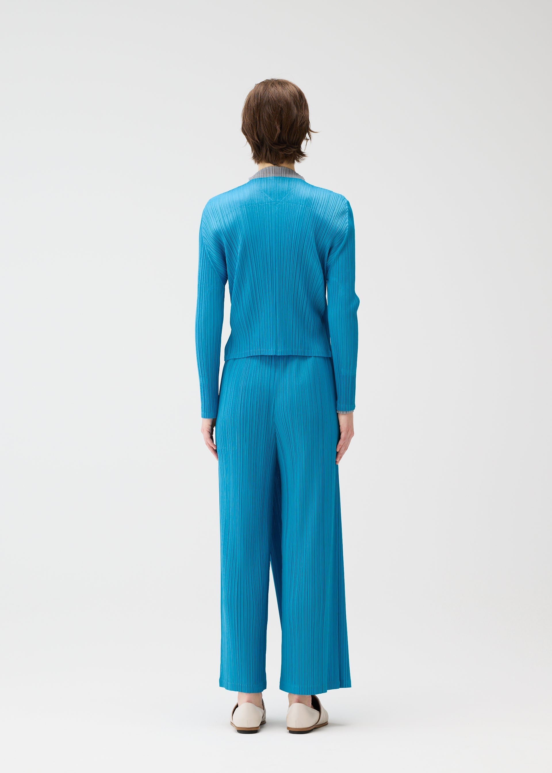 Zoomed view A person with short brown hair and white shoes stands against a plain white background, wearing PLEATS PLEASE ISSEY MIYAKE's FACADE set: a matching bright blue, long-sleeve pleated top with subtle cord design and wide-leg pants.