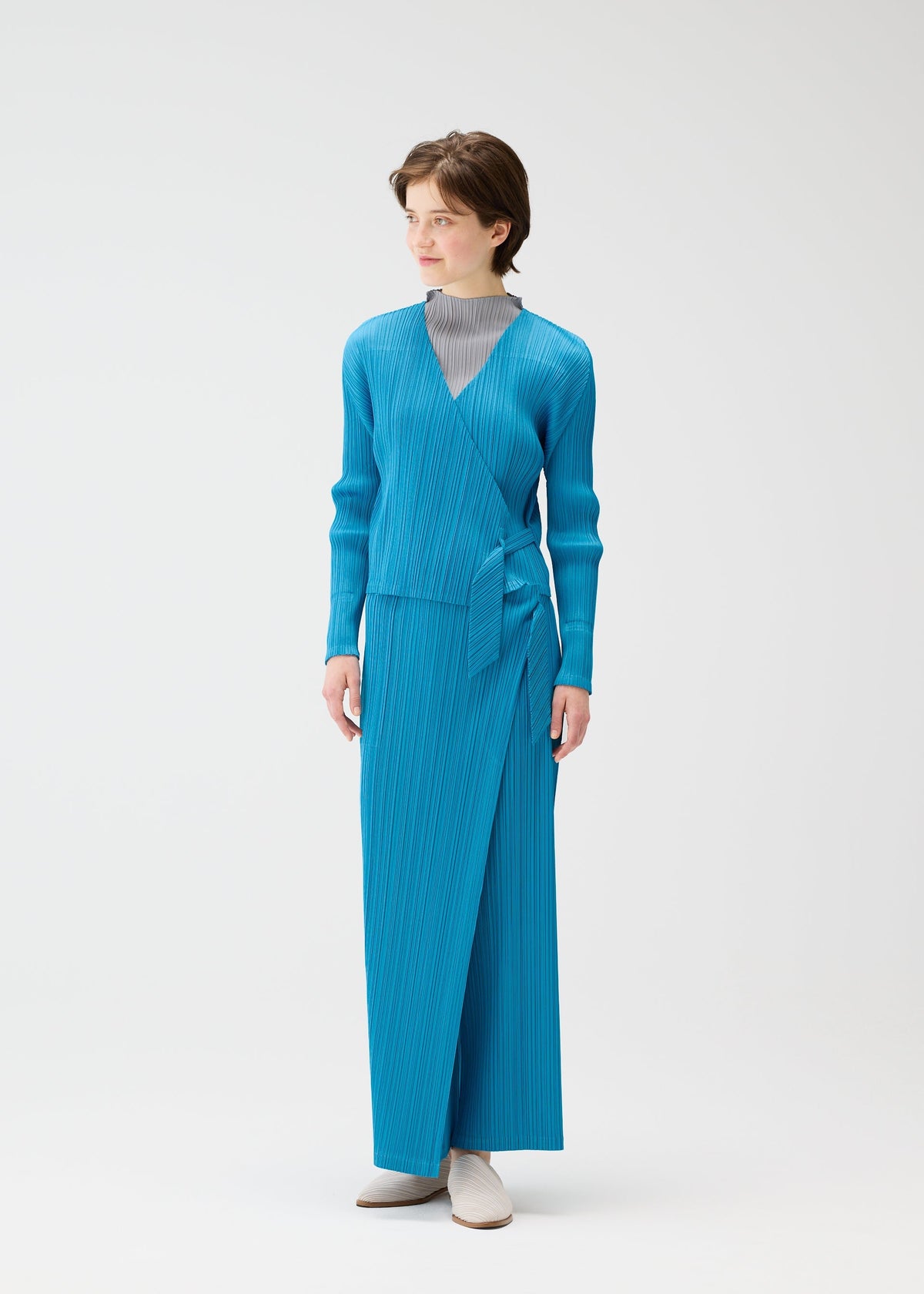 A person wears the PLEATS PLEASE ISSEY MIYAKE FACADE set—a bright blue long-sleeved wrap top with adjustable cords and matching ankle-length skirt—over a light gray high-neck top, paired with white shoes, against a white background. view 9 of 9