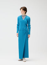 A person wears the PLEATS PLEASE ISSEY MIYAKE FACADE set—a bright blue long-sleeved wrap top with adjustable cords and matching ankle-length skirt—over a light gray high-neck top, paired with white shoes, against a white background.