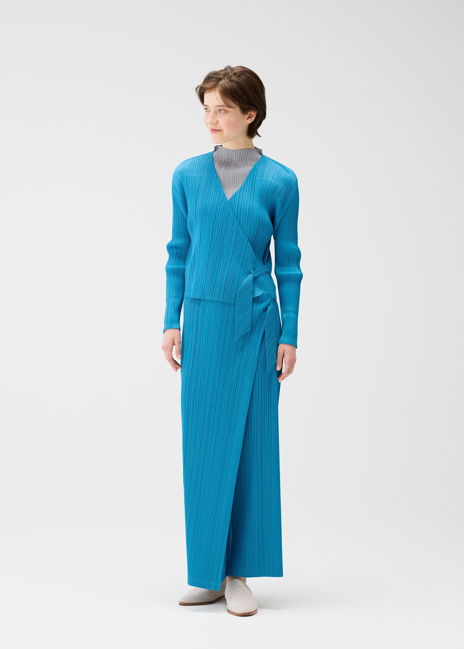 Zoomed view A person wears the PLEATS PLEASE ISSEY MIYAKE FACADE set—a bright blue long-sleeved wrap top with adjustable cords and matching ankle-length skirt—over a light gray high-neck top, paired with white shoes, against a white background.