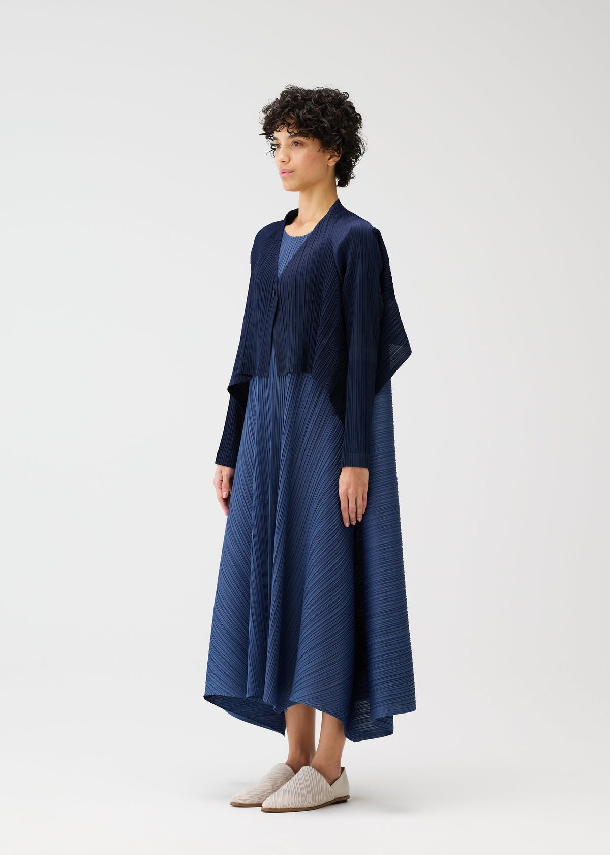 A person with short curly hair models the CITY FRAME navy pleated dress and pleated cardigan by PLEATS PLEASE ISSEY MIYAKE, paired with light slip-on shoes, standing against a plain white background. view 6 of 8