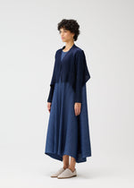 A person with short curly hair models the CITY FRAME navy pleated dress and pleated cardigan by PLEATS PLEASE ISSEY MIYAKE, paired with light slip-on shoes, standing against a plain white background.