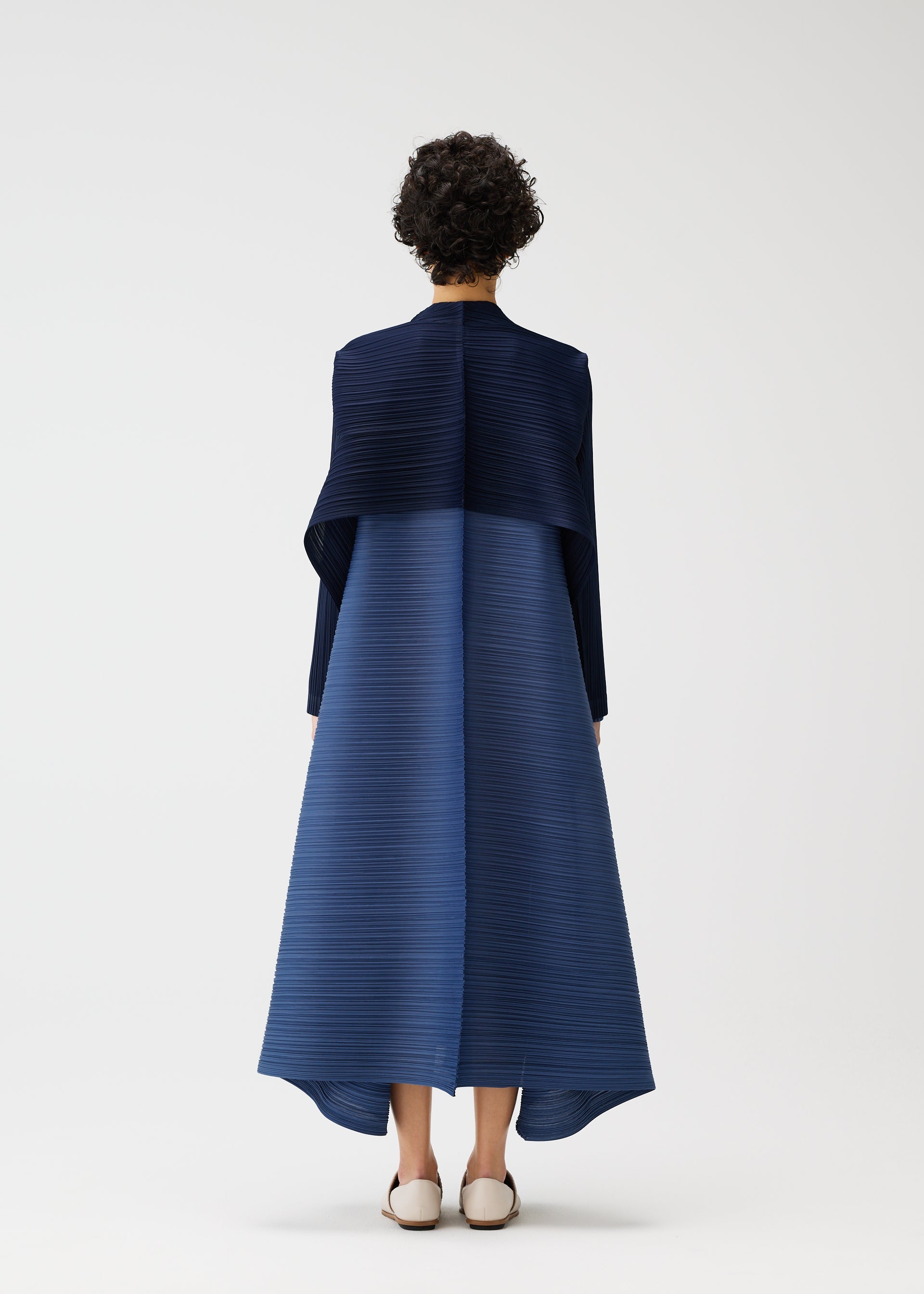 Zoomed view A person with short curly hair, seen from behind, wears the CITY FRAME dress by PLEATS PLEASE ISSEY MIYAKE—an elegant, long-sleeved, pleated navy and blue piece with a voluminous silhouette—paired with white flats against a plain white background.