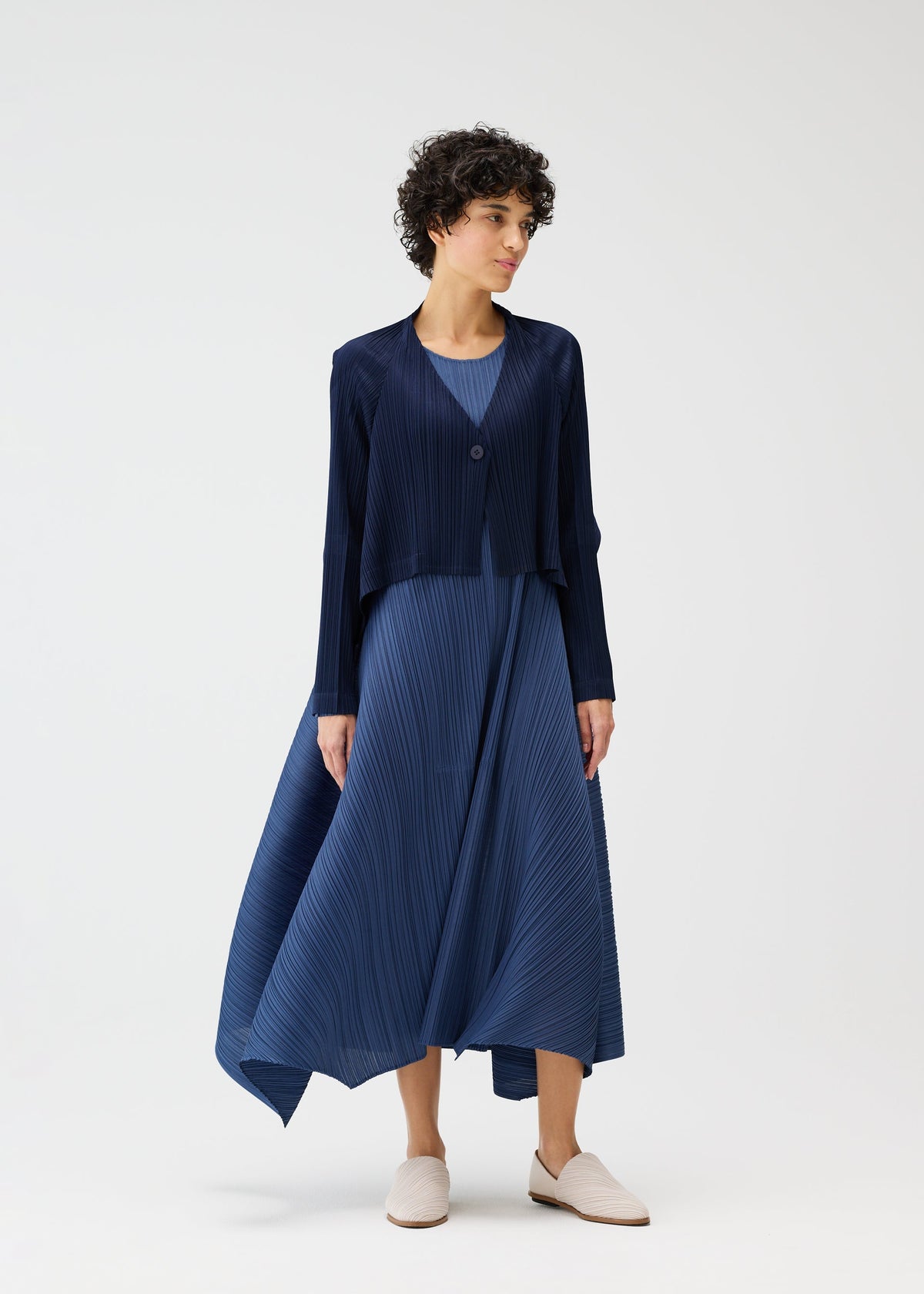 A person wears the CITY FRAME navy pleated dress and matching cardigan by PLEATS PLEASE ISSEY MIYAKE, paired with light slip-on shoes, standing against a plain white background. view 8 of 8