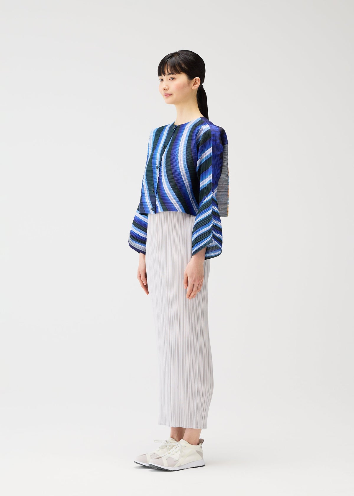 A woman with long dark hair tied back wears the CITY COLLAGE striped blue, purple, and black buttoned cardigan by PLEATS PLEASE ISSEY MIYAKE, a white pleated skirt with horizontal pleating, and white sneakers against a plain white background. view 6 of 7