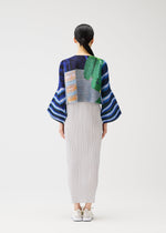 A woman with tied-back dark hair wears the CITY COLLAGE skirt and top by PLEATS PLEASE ISSEY MIYAKE: a long, pleated light skirt and a blue wide-sleeved top with abstract green, blue, and orange patterns. She pairs them with white shoes.