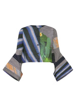 The CITY COLLAGE blouse by PLEATS PLEASE ISSEY MIYAKE features a cropped, long-sleeve design with flared cuffs, front buttons, and horizontal pleats. It showcases blue, green, gray, and orange collage patterns with stripes and abstract florals. - GRAY×NAVY （no.09）