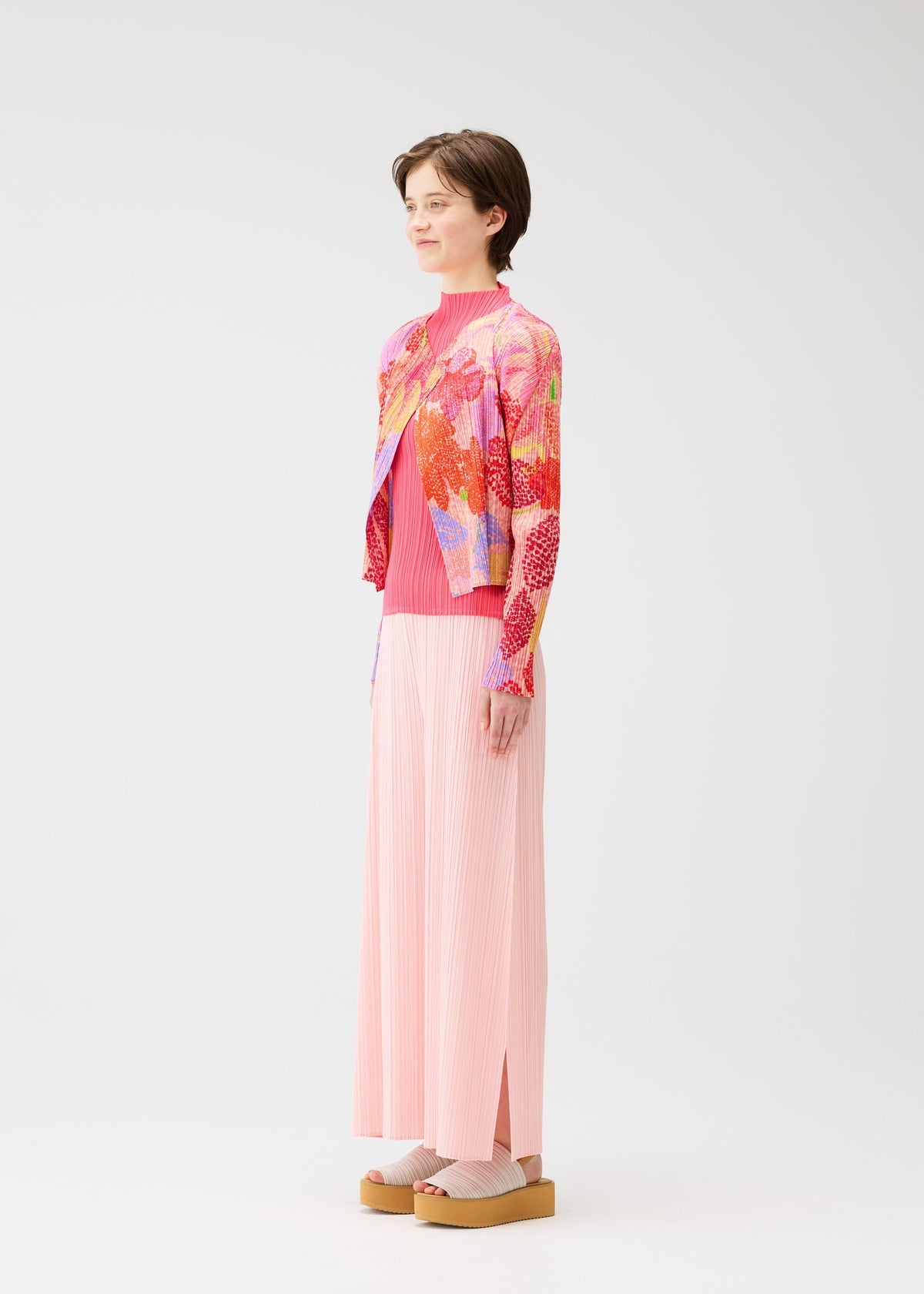 A person stands sideways wearing the vibrant "FOGGY FOREST" cardigan by PLEATS PLEASE ISSEY MIYAKE, paired with a pink pleated top, a long pink pleated skirt, and beige platform sandals against a plain white background. view 5 of 6