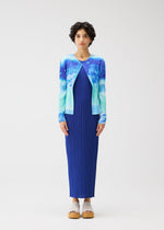 A person with short, curly hair stands against a plain background, wearing the SEA BUBBLE pleated maxi dress by PLEATS PLEASE ISSEY MIYAKE and a blue-and-teal tie-dye cardigan, paired with beige platform sandals.