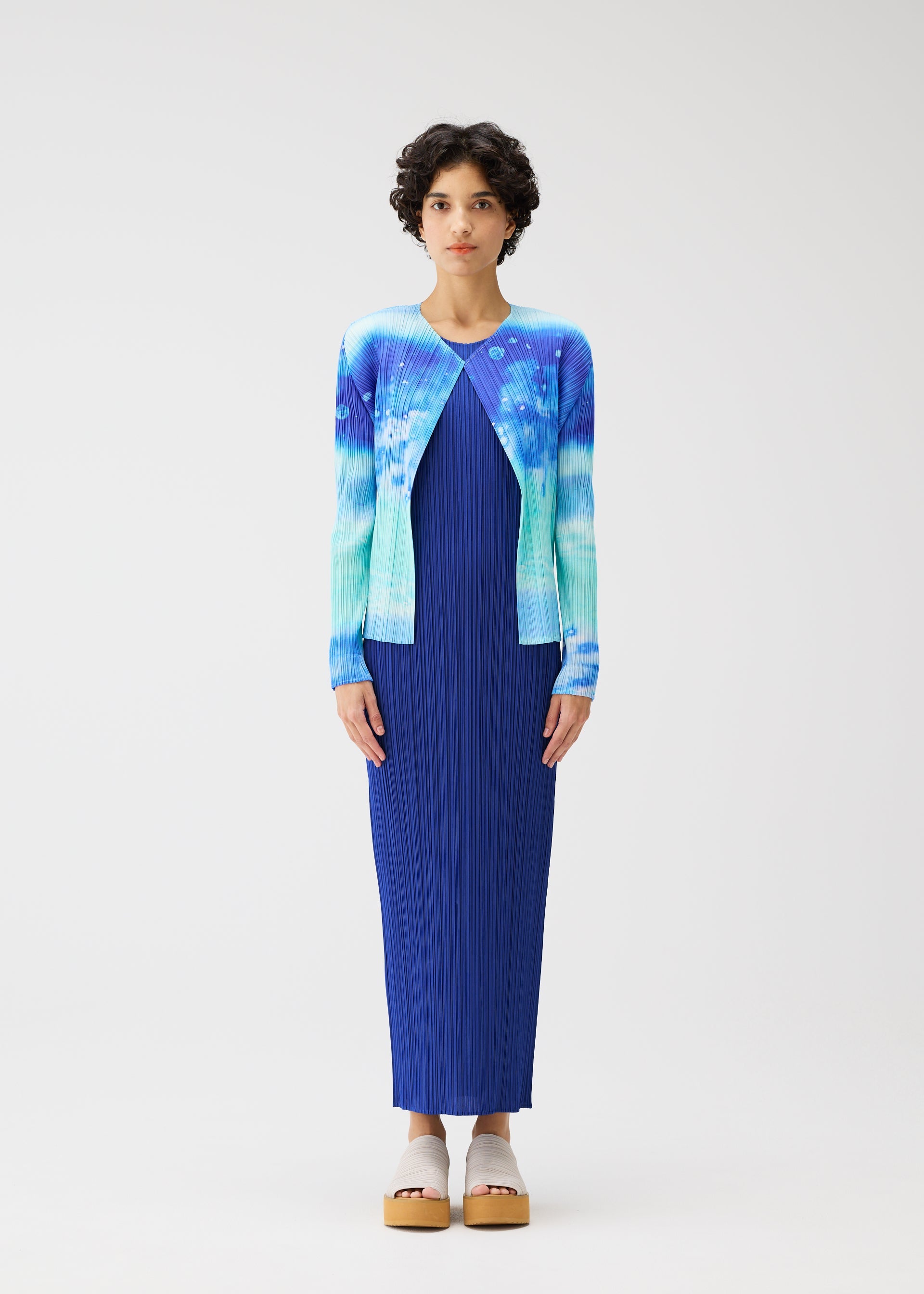 Zoomed view A person with short, curly hair stands against a plain background, wearing the SEA BUBBLE pleated maxi dress by PLEATS PLEASE ISSEY MIYAKE and a blue-and-teal tie-dye cardigan, paired with beige platform sandals.
