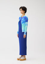 A person in profile stands against a plain background, wearing the SEA BUBBLE blue pleated dress from PLEATS PLEASE ISSEY MIYAKE, paired with a sea-inspired cardigan and beige platform sandals. They have short curly hair.