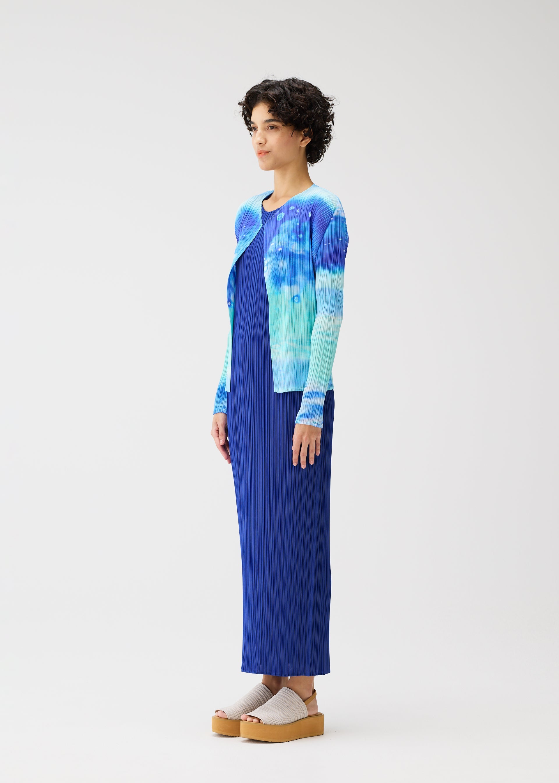 Zoomed view A person in profile stands against a plain background, wearing the SEA BUBBLE blue pleated dress from PLEATS PLEASE ISSEY MIYAKE, paired with a sea-inspired cardigan and beige platform sandals. They have short curly hair.