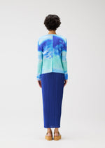 A person with short curly hair wears the SEA BUBBLE blue tie-dye long-sleeve top and pleated skirt by PLEATS PLEASE ISSEY MIYAKE, paired with tan platform sandals, standing against a plain white background.