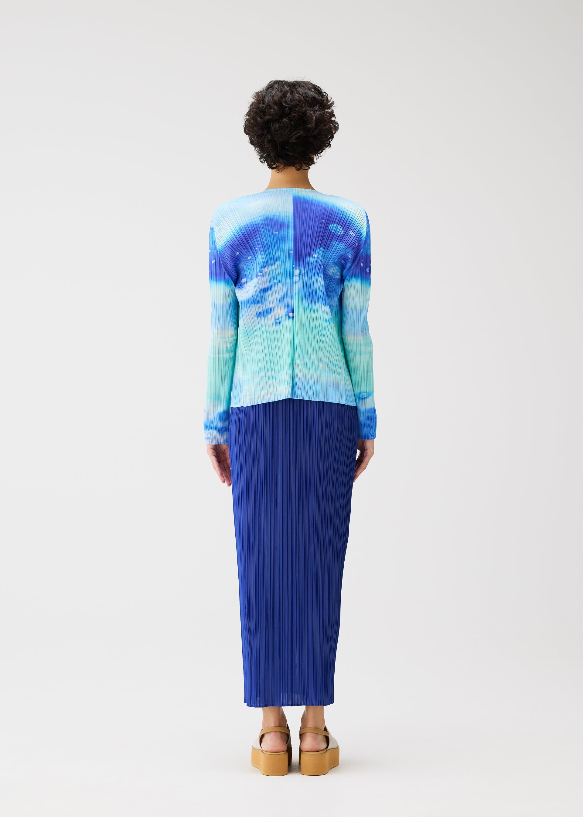 Zoomed view A person with short curly hair wears the SEA BUBBLE blue tie-dye long-sleeve top and pleated skirt by PLEATS PLEASE ISSEY MIYAKE, paired with tan platform sandals, standing against a plain white background.