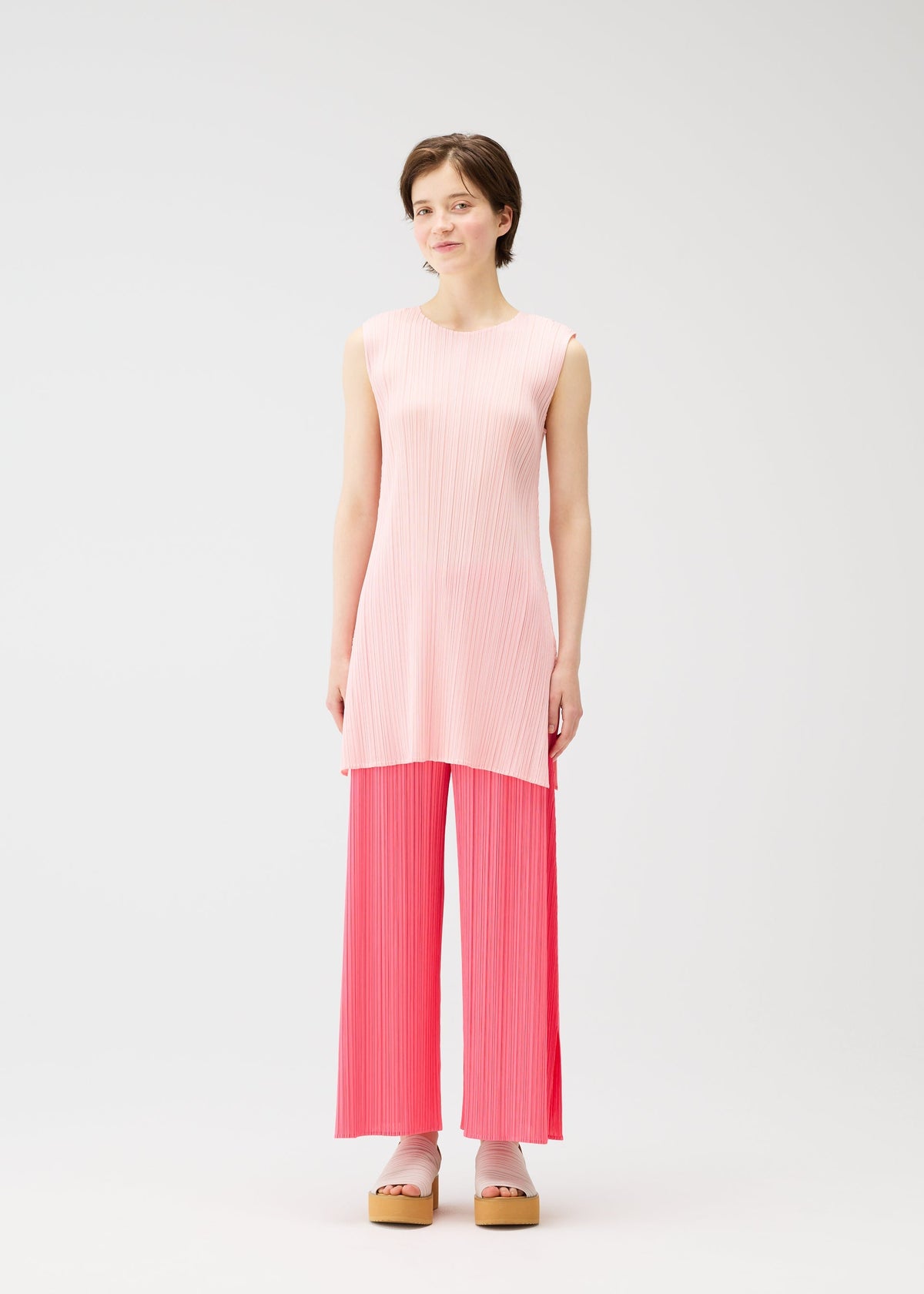 A person with short brown hair wears MONTHLY COLORS : MARCH by PLEATS PLEASE ISSEY MIYAKE—a sleeveless, round-neck tunic in light pink and bright pink pleated wide-leg pants—styled with platform sandals, standing against a plain white background. view 8 of 11