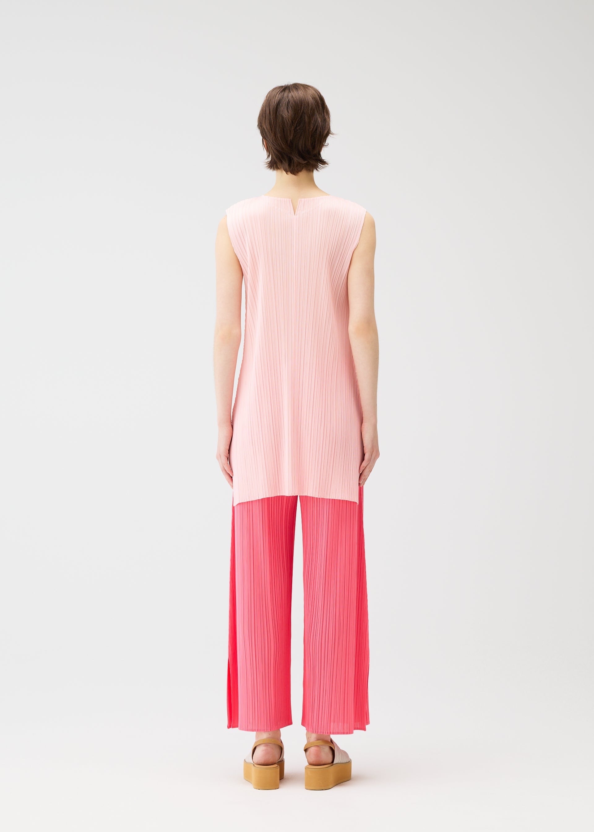 Zoomed view Someone stands with their back to the camera in a light pink sleeveless tunic and bright pink pleated wide-leg pants from MONTHLY COLORS : MARCH by PLEATS PLEASE ISSEY MIYAKE, styled with platform sandals, short brown hair, and a plain white background.