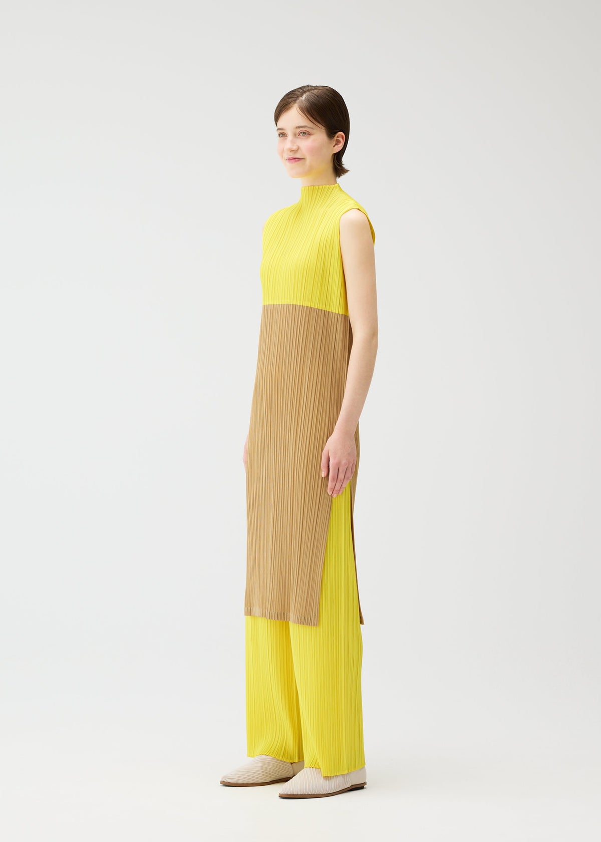 In a studio, a person with short brown hair wears the SWING tunic from PLEATS PLEASE ISSEY MIYAKE—a high-neck, sleeveless two-tone yellow top and pants with a light brown panel—looking slightly to the side with a neutral expression. view 8 of 12