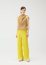 A woman stands smiling against a white background, wearing the tan high-neck sleeveless SWING tunic and wide-leg yellow pleated pants from PLEATS PLEASE ISSEY MIYAKE, paired with cream shoes. Her straight brown hair is pulled back for a versatile look.