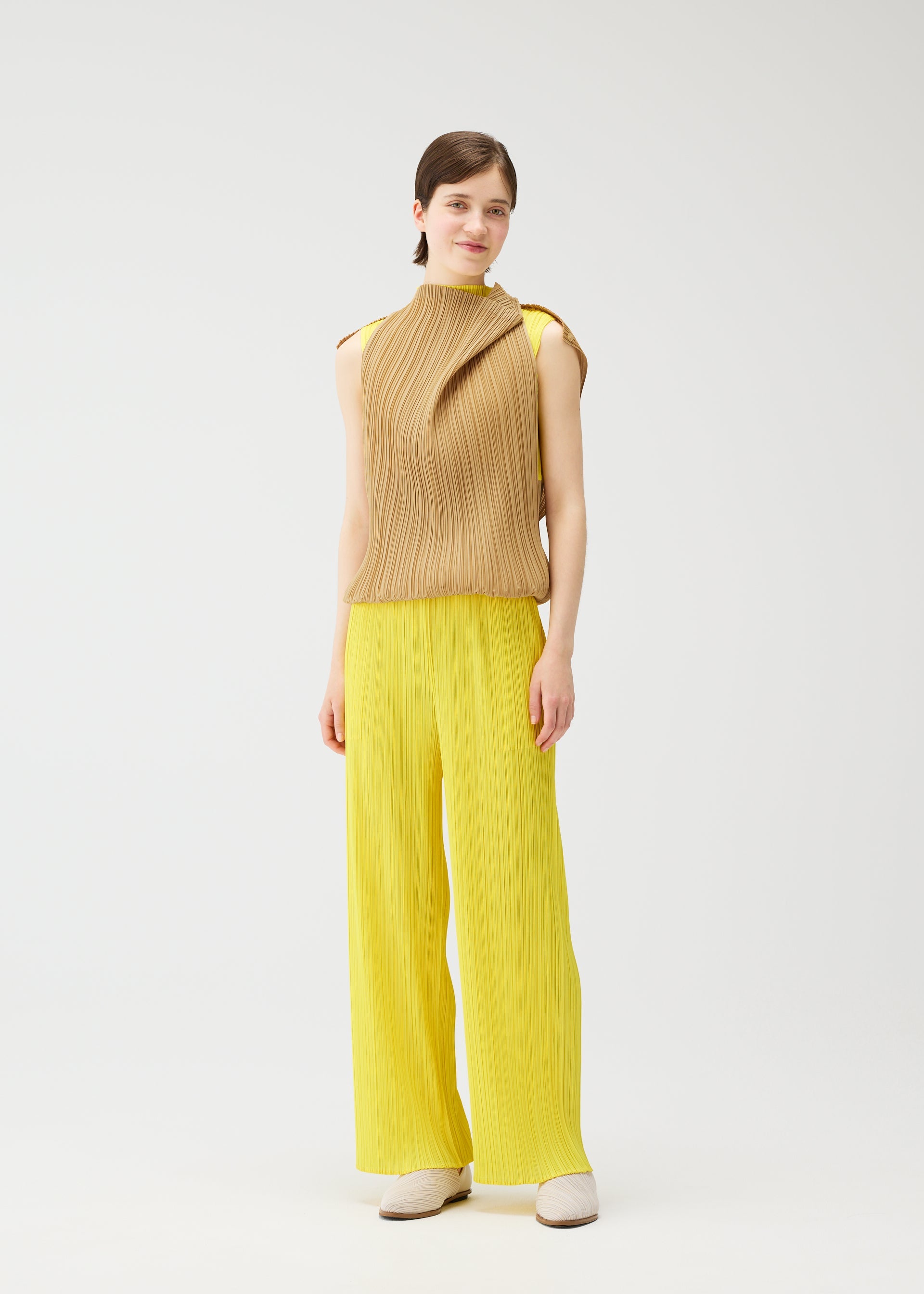 Zoomed view A woman stands smiling against a white background, wearing the tan high-neck sleeveless SWING tunic and wide-leg yellow pleated pants from PLEATS PLEASE ISSEY MIYAKE, paired with cream shoes. Her straight brown hair is pulled back for a versatile look.