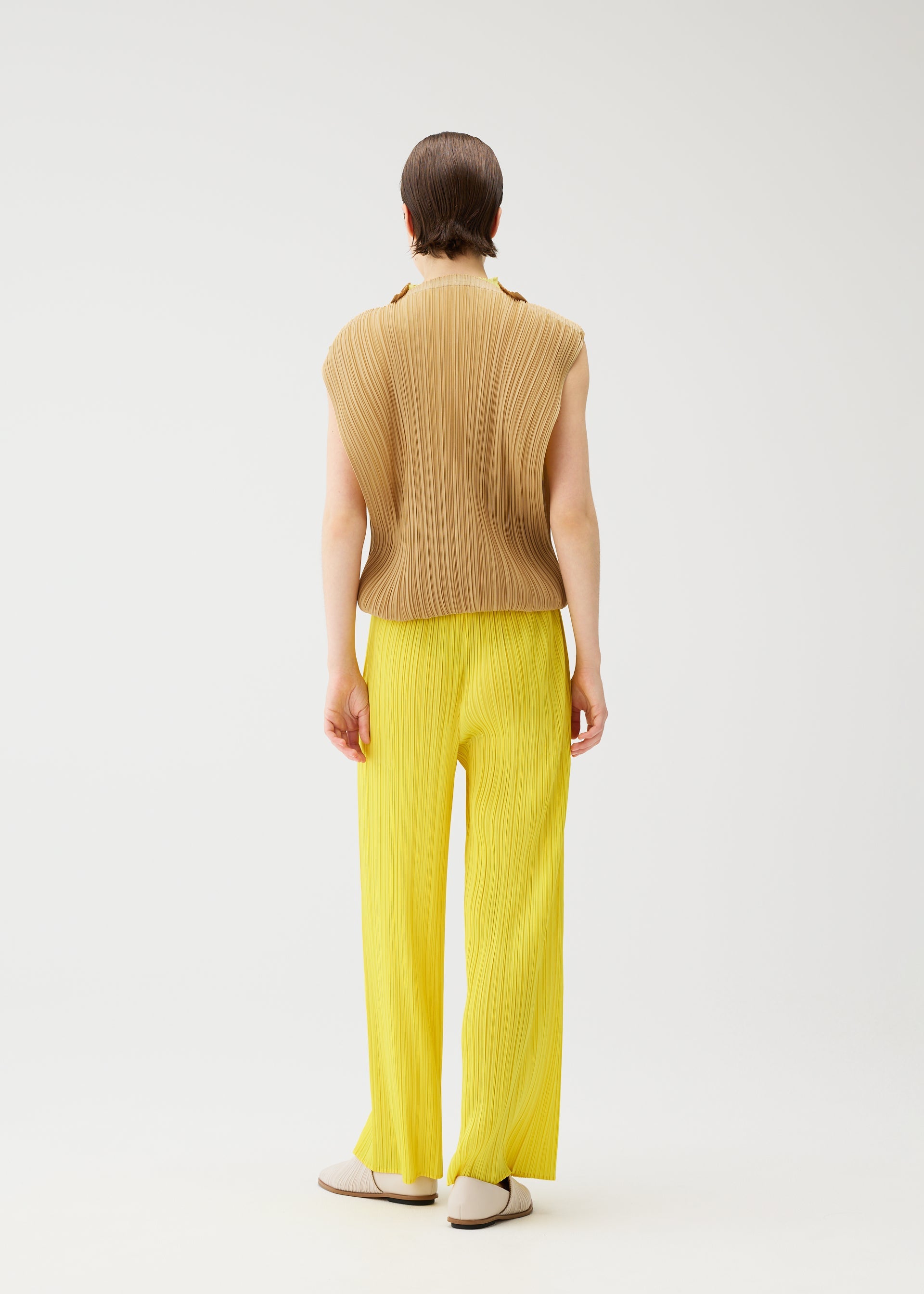Zoomed view A person faces away in the SWING tunic by PLEATS PLEASE ISSEY MIYAKE, styled in brown with bright yellow pleated pants and white shoes, highlighting versatile looks against a plain white background.
