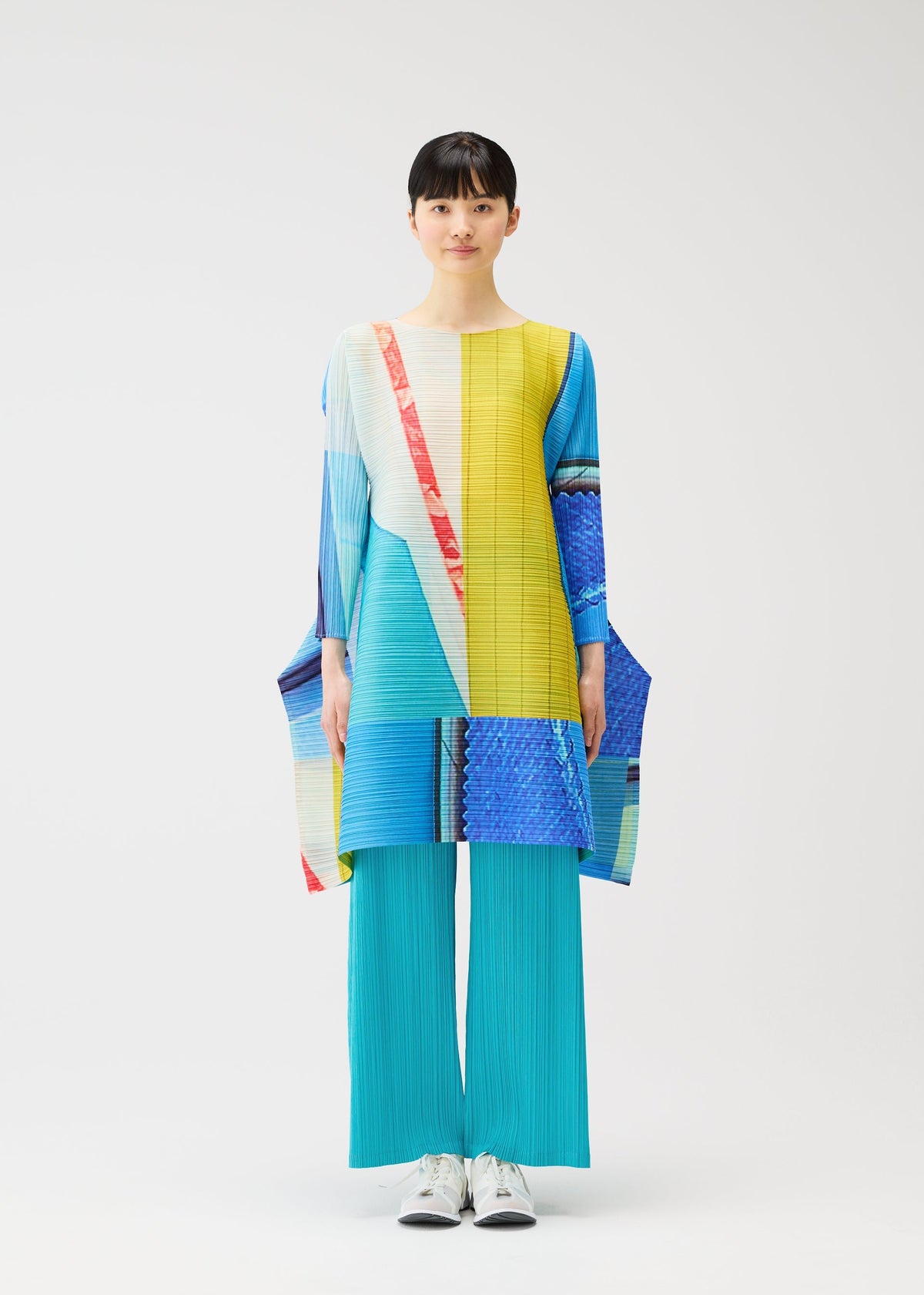 A person stands facing forward against a plain white background, wearing PLEATS PLEASE ISSEY MIYAKE’s CITY COLLAGE: turquoise pleated pants and a colorful, long-sleeved tunic with yellow, blue, red, and white geometric patterns. view 6 of 9