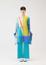 A person stands facing forward against a plain white background, wearing PLEATS PLEASE ISSEY MIYAKE’s CITY COLLAGE: turquoise pleated pants and a colorful, long-sleeved tunic with yellow, blue, red, and white geometric patterns.