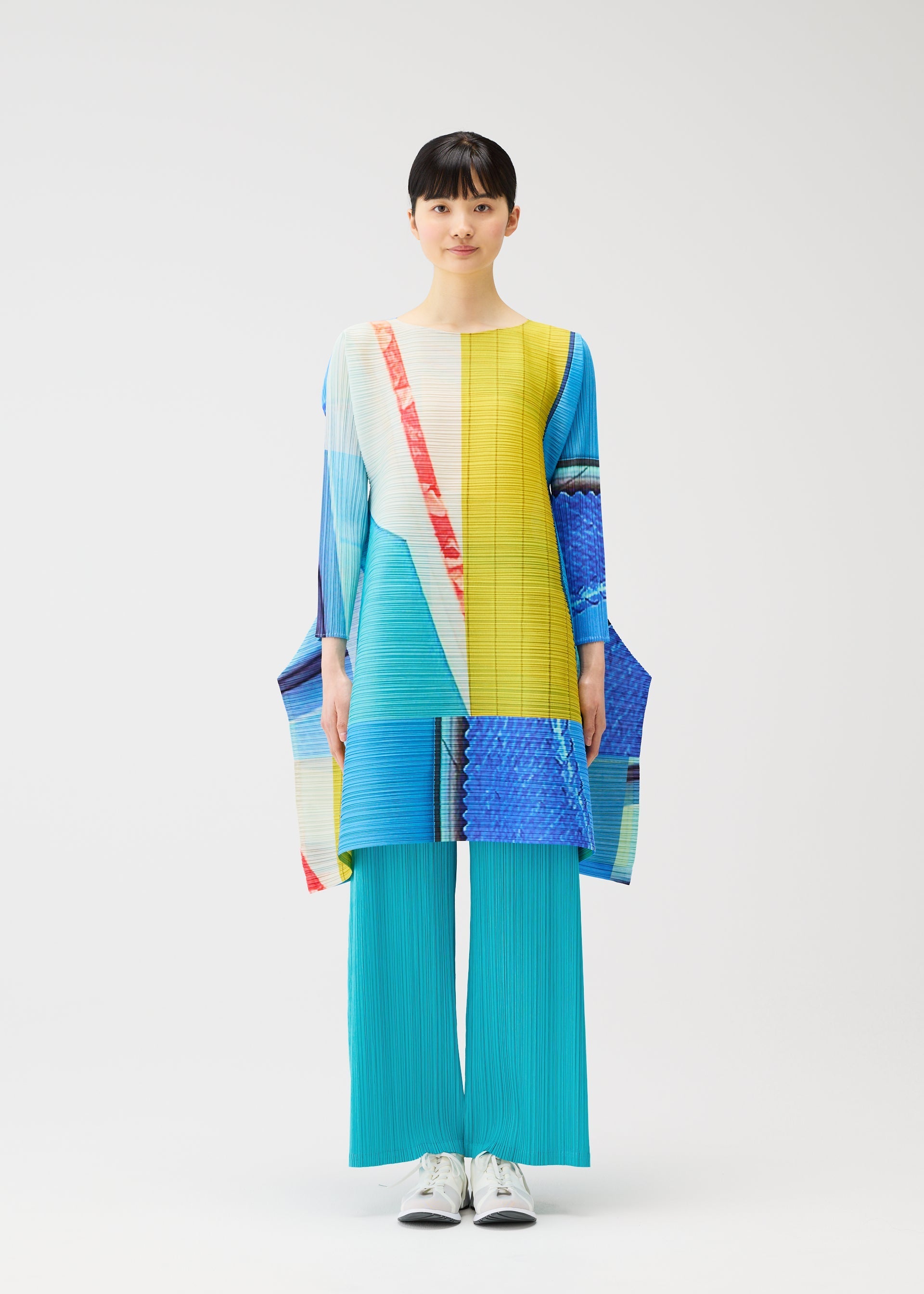 Zoomed view A person stands facing forward against a plain white background, wearing PLEATS PLEASE ISSEY MIYAKE’s CITY COLLAGE: turquoise pleated pants and a colorful, long-sleeved tunic with yellow, blue, red, and white geometric patterns.