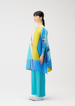 A woman stands in profile wearing the CITY COLLAGE long-sleeved tunic in blue, yellow, and red by PLEATS PLEASE ISSEY MIYAKE, paired with loose turquoise pants and white shoes against a plain white background.