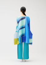 A person stands with their back to the camera, wearing the CITY COLLAGE tunic in blue, yellow, and aqua stripes with wide turquoise pleated pants by PLEATS PLEASE ISSEY MIYAKE. Their hair is tied back against a plain white background.