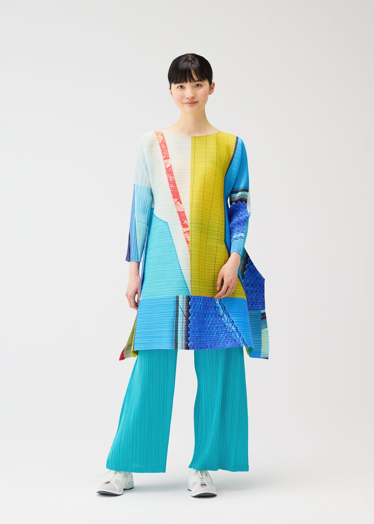 A person wears the CITY COLLAGE tunic by PLEATS PLEASE ISSEY MIYAKE, featuring geometric blue, yellow, and red patterns, paired with bright turquoise pleated pants and white sneakers against a plain white background. view 9 of 9