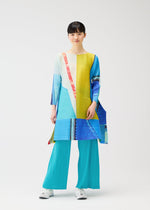 A person wears the CITY COLLAGE tunic by PLEATS PLEASE ISSEY MIYAKE, featuring geometric blue, yellow, and red patterns, paired with bright turquoise pleated pants and white sneakers against a plain white background.