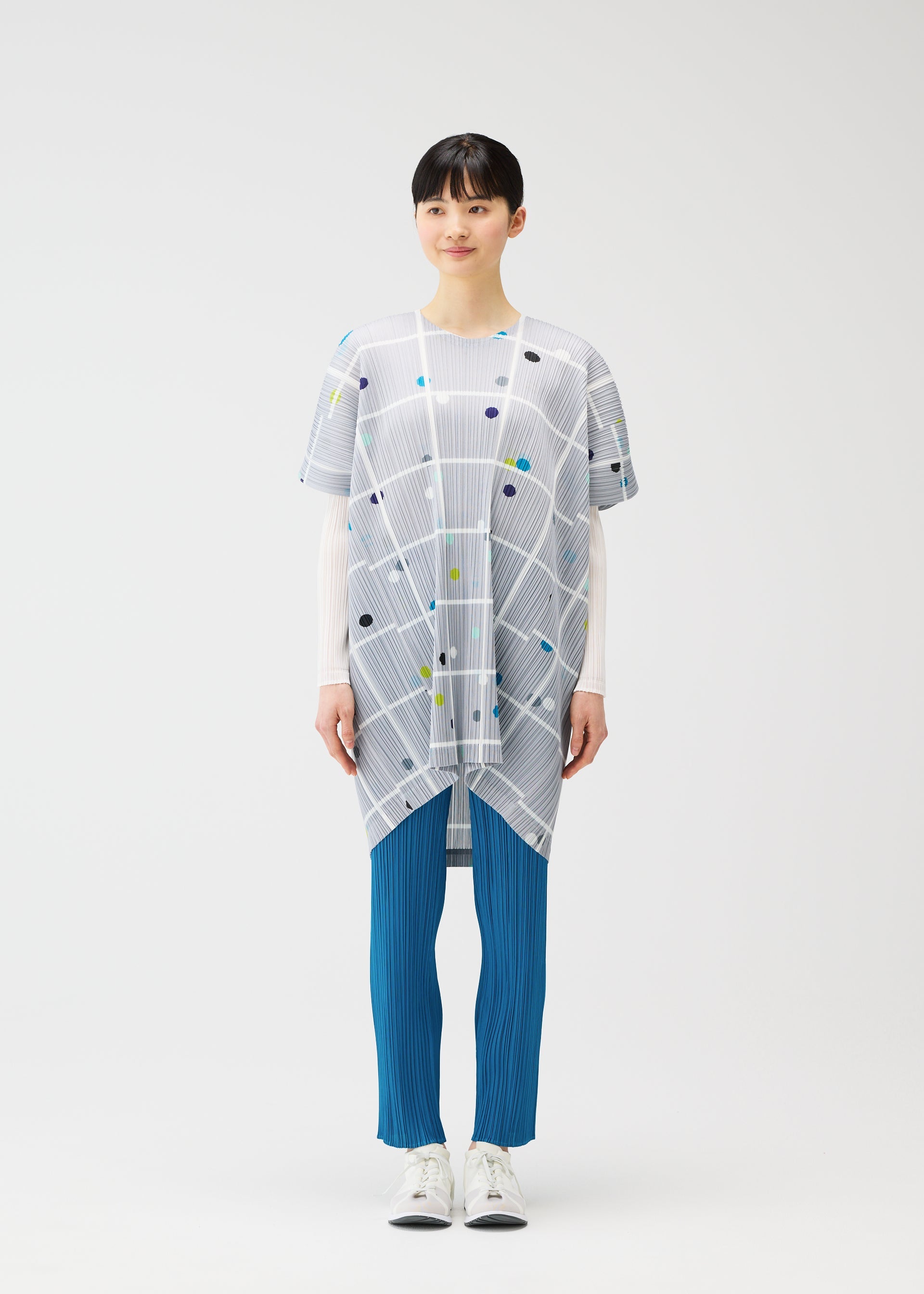Zoomed view A person faces the camera wearing the PLEATS PLEASE ISSEY MIYAKE CROSSING DOTS tunic over a long-sleeve white top, blue pleated pants, and white sneakers, against a plain white background.