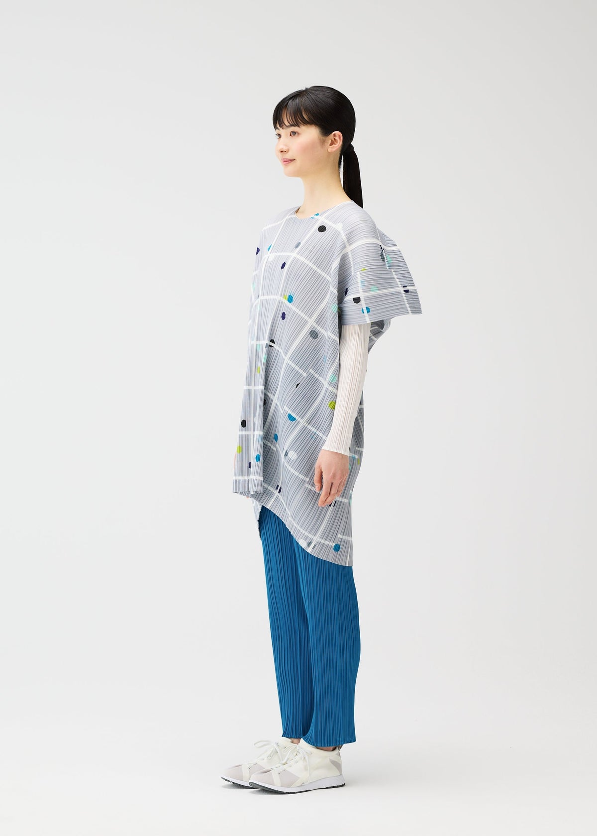 A woman stands in profile against a white background, wearing the PLEATS PLEASE ISSEY MIYAKE CROSSING DOTS light gray round-neck tunic with a plaid and dots pattern, layered over a white top, blue pleated pants, and white sneakers. view 7 of 9
