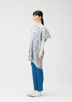 A woman stands in profile against a white background, wearing the PLEATS PLEASE ISSEY MIYAKE CROSSING DOTS light gray round-neck tunic with a plaid and dots pattern, layered over a white top, blue pleated pants, and white sneakers.