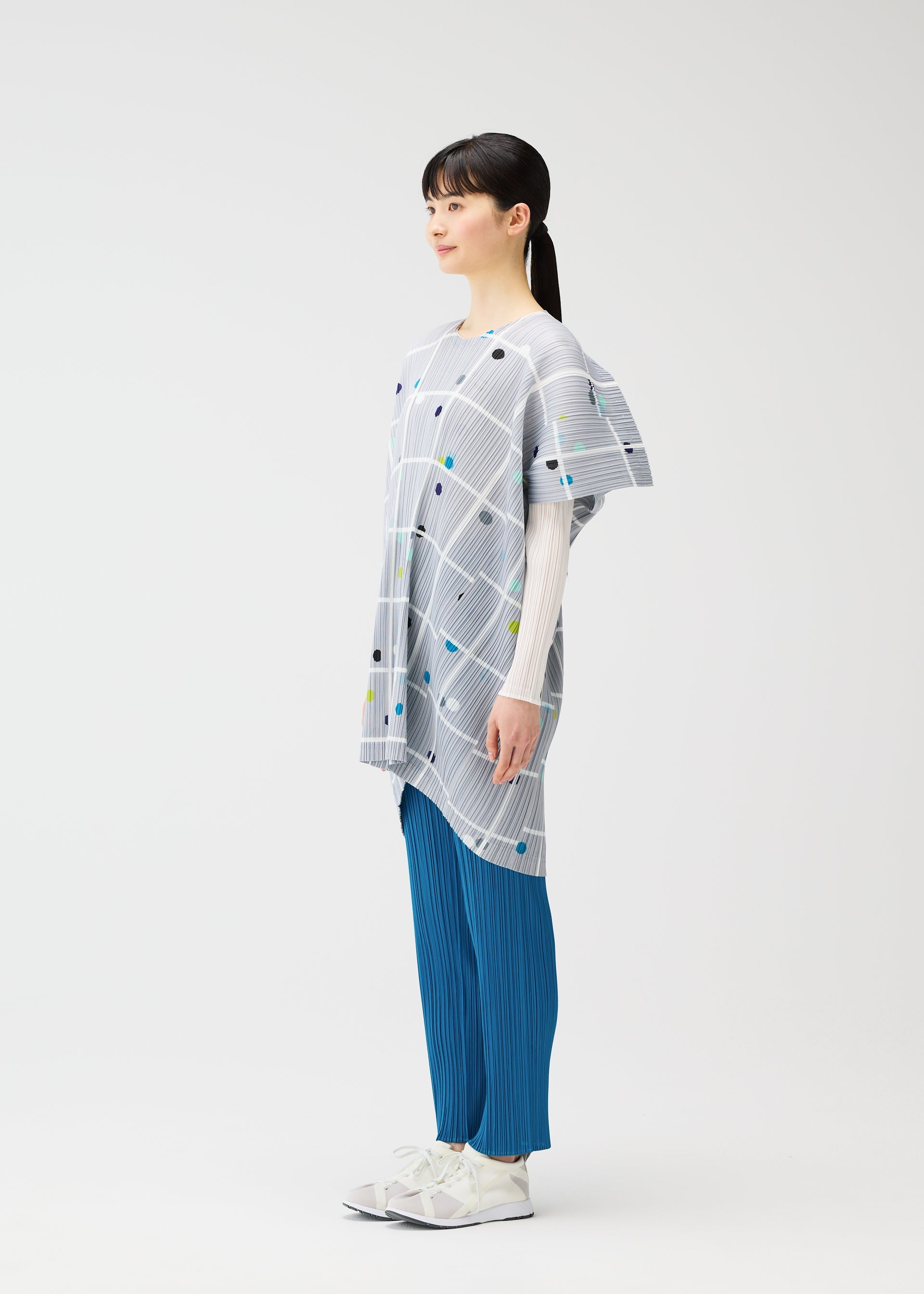 Zoomed view A woman stands in profile against a white background, wearing the PLEATS PLEASE ISSEY MIYAKE CROSSING DOTS light gray round-neck tunic with a plaid and dots pattern, layered over a white top, blue pleated pants, and white sneakers.